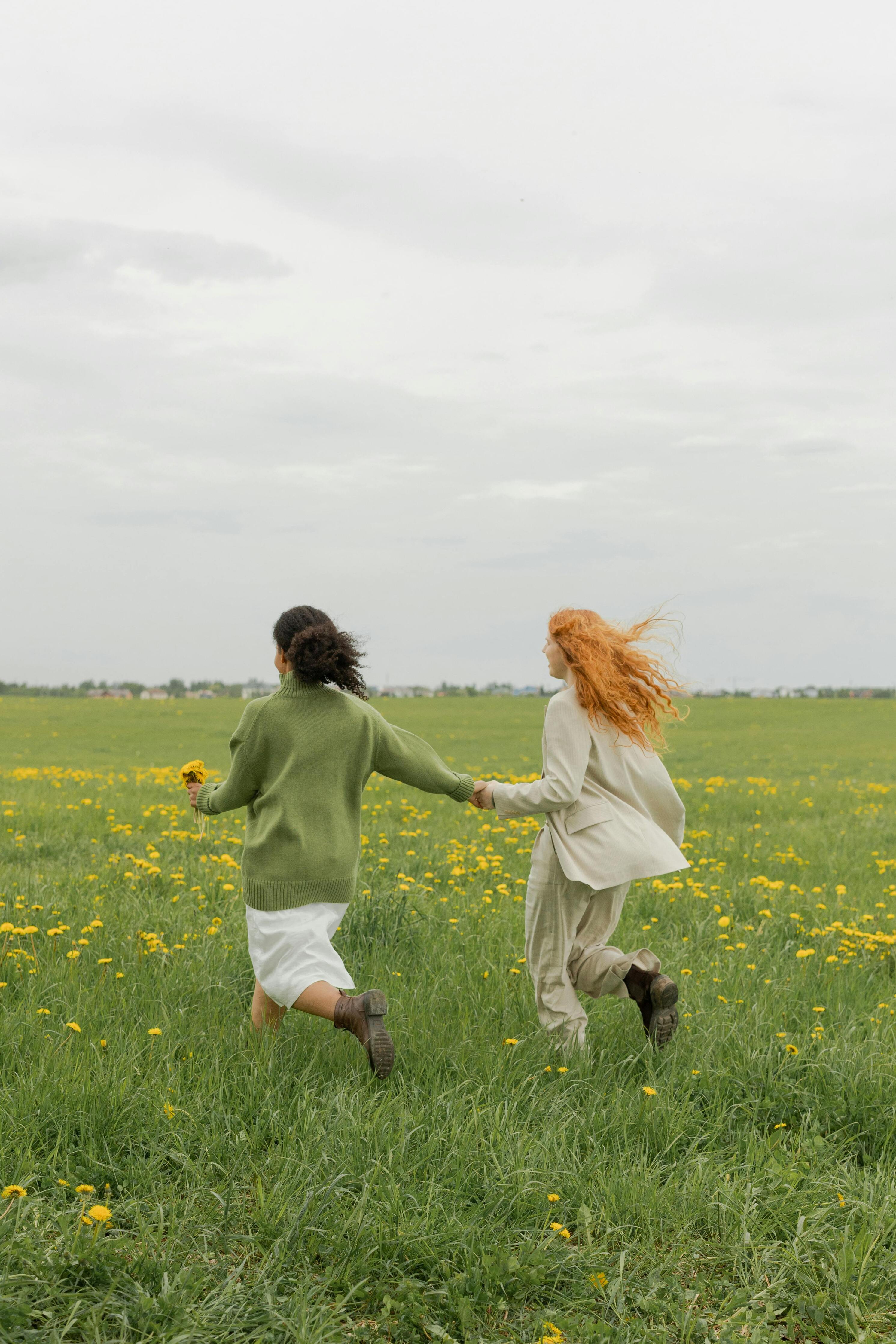 Two women running hand in hand through a meadow with yellow wildflowers, their hair flowing in the wind, evoking a sense of freedom and tranquility.