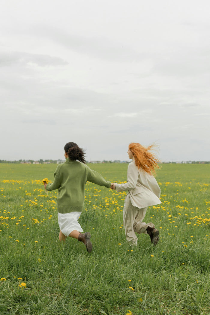 Two women running hand in hand through a meadow with yellow wildflowers, their hair flowing in the wind, evoking a sense of freedom and tranquility.